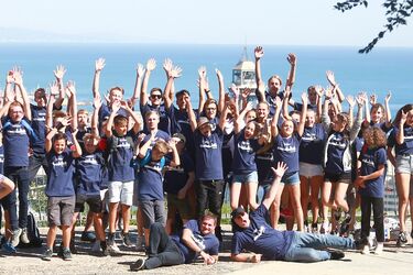 eine Gruppe Jugendlicher reißt begeistert die Hände in die Luft, im Hintergrund das Meer bei strahlendem Sonnenschein
